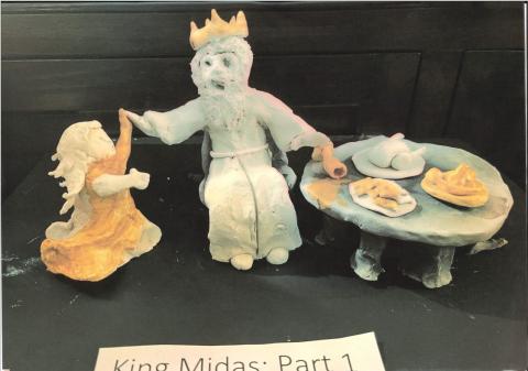 A clay model of Midas at the dining table eating food that is beginning to turn to gold. His daughter has just run to him and is in the process of turning to gold.